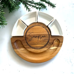 May include: A wooden serving platter with a circular design, featuring a central engraved circle with "The Sampson Family" and five white ceramic bowls. The platter has a natural wood grain finish and is ideal for serving appetizers.
