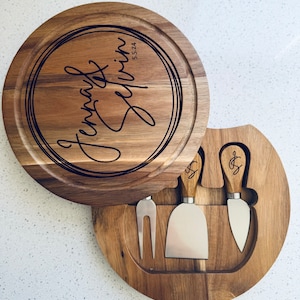 May include: A round wooden cheese board with a lid that opens to reveal a set of three cheese knives. The board is engraved with the text "Jenna & Griffin 5.5.24".