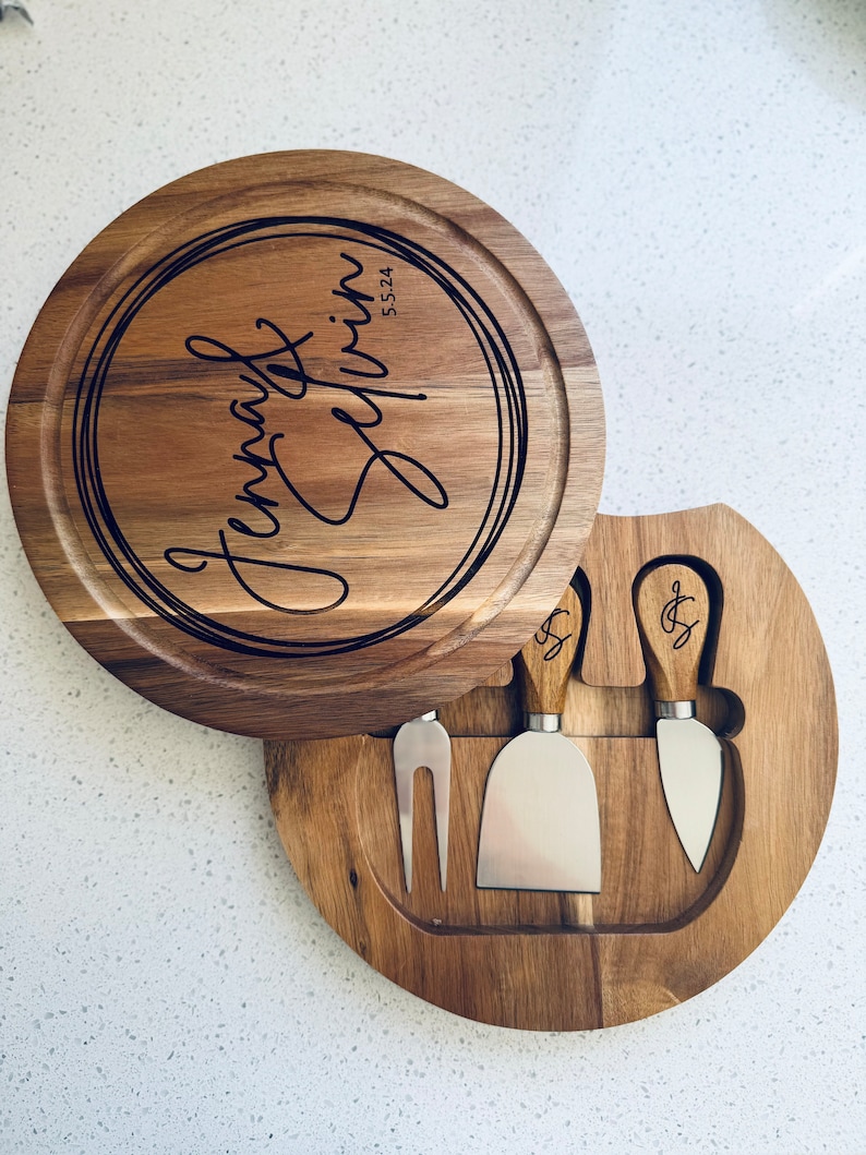 May include: A round wooden cheese board with a lid that opens to reveal a set of three cheese knives. The board is engraved with the text "Jenna & Griffin 5.5.24".