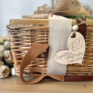 May include: A wicker picnic basket with a brown leather handle and a wooden heart-shaped tag that says "Congratulations and best wishes!" with the initials "B&T" and the text "XOX0" in the center of the heart. The tag is attached to the basket with a brown leather strap and a white wooden bead.