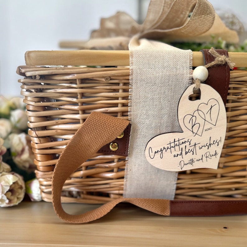 May include: A wicker picnic basket with a brown leather handle and a wooden heart-shaped tag that says "Congratulations and best wishes!" with the initials "B&T" and the text "XOX0" in the center of the heart. The tag is attached to the basket with a brown leather strap and a white wooden bead.