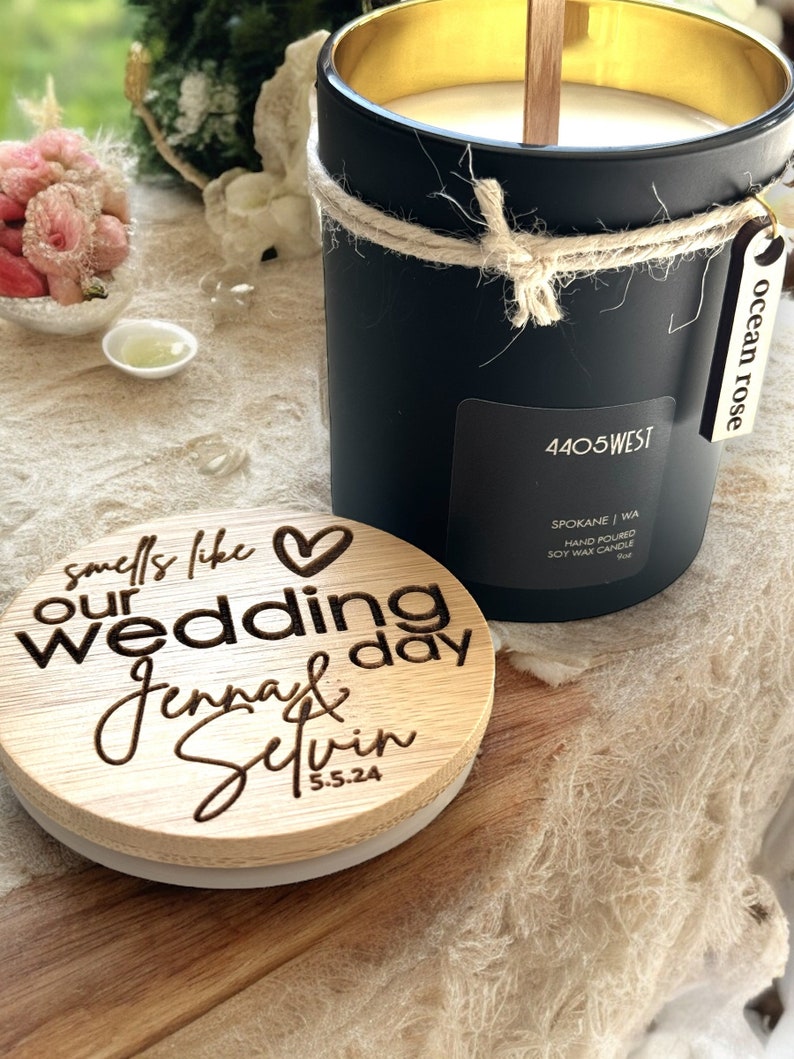 May include: A black soy wax candle with a gold lid and a wooden wick. The candle is labeled "4405 WEST" and "ocean rose". The candle is packaged with a wooden lid that says "smells like our wedding day Jenna & Selvin 5.5.24".