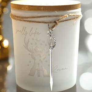 May include: A white candle with a wooden lid and a decorative sword charm. The candle is wrapped with twine and features the text "smells like" and "Rowan", along with a deer and sword graphic.