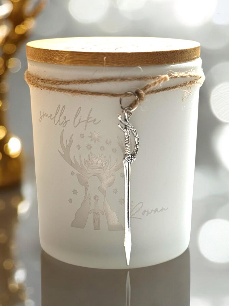 May include: A white candle with a wooden lid and a decorative sword charm. The candle is wrapped with twine and features the text "smells like" and "Rowan", along with a deer and sword graphic.