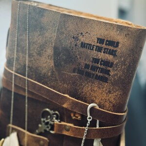 May include: A brown leather journal with the quote "You could rattle the stars, you could do anything if you only dared" printed on the cover. The journal is secured with a leather strap and adorned with decorative charms.
