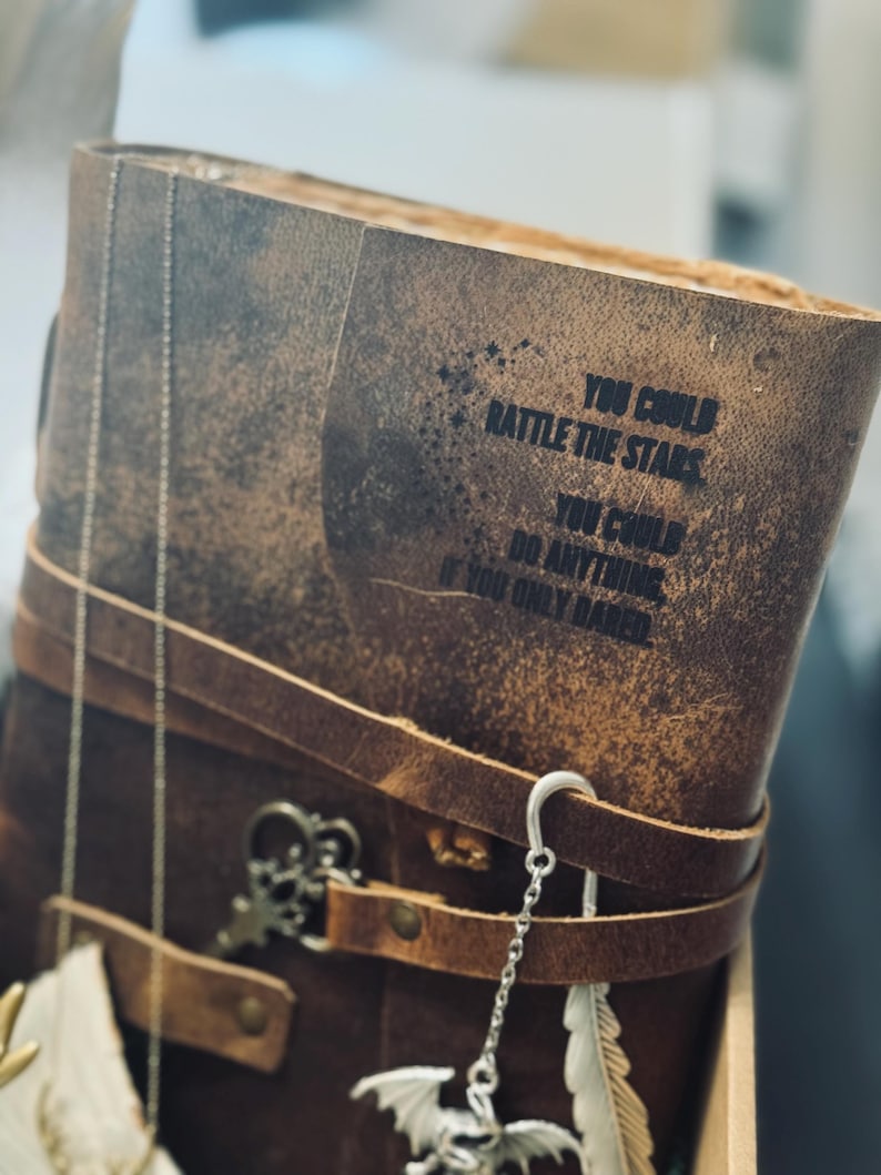 May include: A brown leather journal with the quote "You could rattle the stars, you could do anything if you only dared" printed on the cover. The journal is secured with a leather strap and adorned with decorative charms.