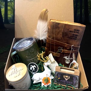 May include: A gift box filled with various items, including a green travel mug, a leather-bound book with text, a white deer figurine, a gold feather, and a candle. The box also contains stickers and a bar of lavender soap.
