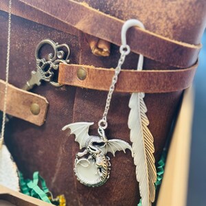 May include: A brown leather journal with a silver dragon charm bookmark and a feather bookmark. A decorative key charm is also visible. The journal is secured with leather straps and metal rivets. The dragon charm has white wings and a glowing orb.
