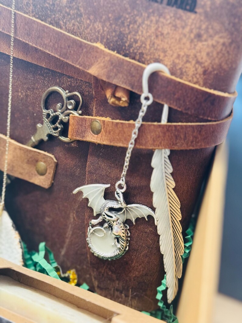 May include: A brown leather journal with a silver dragon charm bookmark and a feather bookmark. A decorative key charm is also visible. The journal is secured with leather straps and metal rivets. The dragon charm has white wings and a glowing orb.