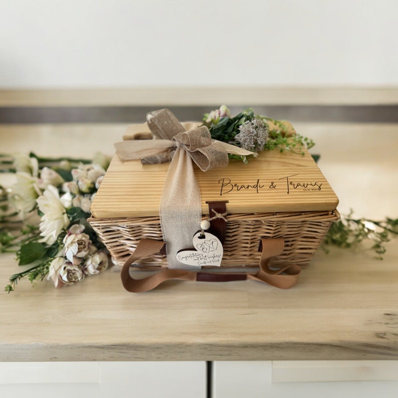 May include: A wicker basket with a wooden lid, personalized with the names "Brandi & Travis" and a heart-shaped tag that reads "Congratulations!" The basket is tied with a brown ribbon and decorated with a bouquet of dried flowers.