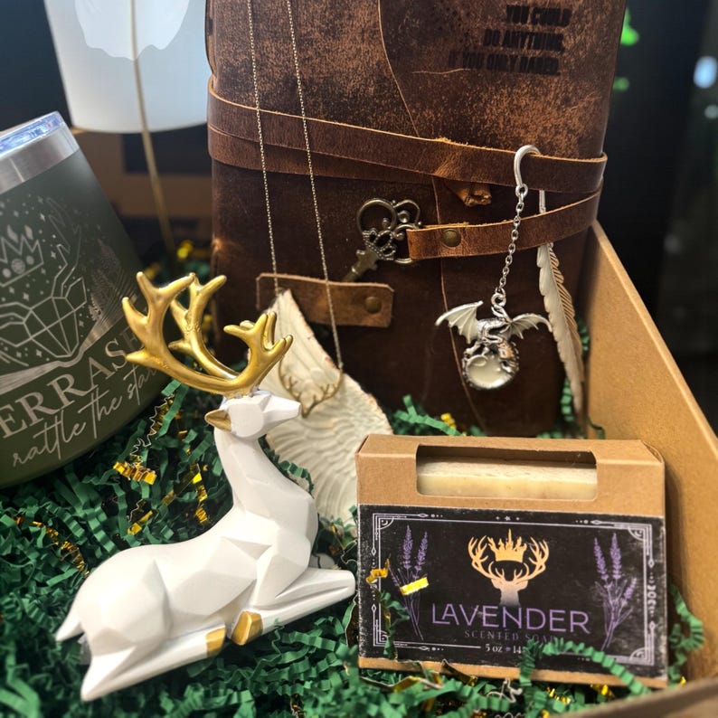 May include: A gift box filled with various items, including a white deer figurine with gold antlers, a leather-bound journal, a lavender-scented soap, and a silver dragon pendant. The box is lined with green shredded paper.