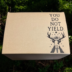 May include: A rectangular cardboard box with the text "YOU DO NOT YIELD" and a black graphic of a stag with a crown and sword. The box is set against a backdrop of green moss.
