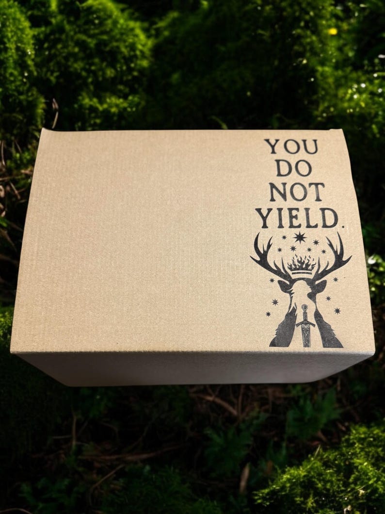 May include: A rectangular cardboard box with the text "YOU DO NOT YIELD" and a black graphic of a stag with a crown and sword. The box is set against a backdrop of green moss.