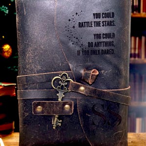 May include: A dark brown leather journal with a strap closure and a decorative key. The journal has the text "YOU COULD RATTLE THE STARS. YOU COULD DO ANYTHING, IF YOU ONLY DARED." embossed on the cover. The initials "SS" are also visible.