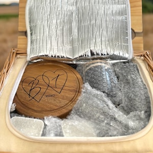 May include: A wicker picnic basket with a wooden cutting board engraved with two hearts and the initials "B" and "T" and the text "XOXO". The basket is filled with bubble wrap and a gray insulated cooler bag.