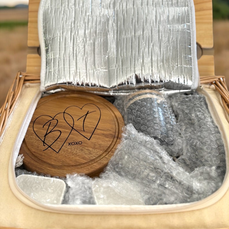 May include: A wicker picnic basket with a wooden cutting board engraved with two hearts and the initials "B" and "T" and the text "XOXO". The basket is filled with bubble wrap and a gray insulated cooler bag.