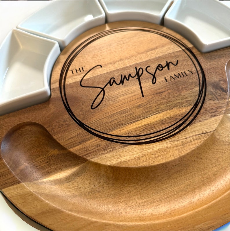 K&ouml;nnte beinhalten: Ein h&ouml;lzernes Serviertablett mit kreisf&ouml;rmigem Design, mit einem zentralen gravierten Bereich mit dem Text "The Sampson Family". Es enth&auml;lt sechs wei&szlig;e Keramikschalen und einen gebogenen Bereich zum Servieren.