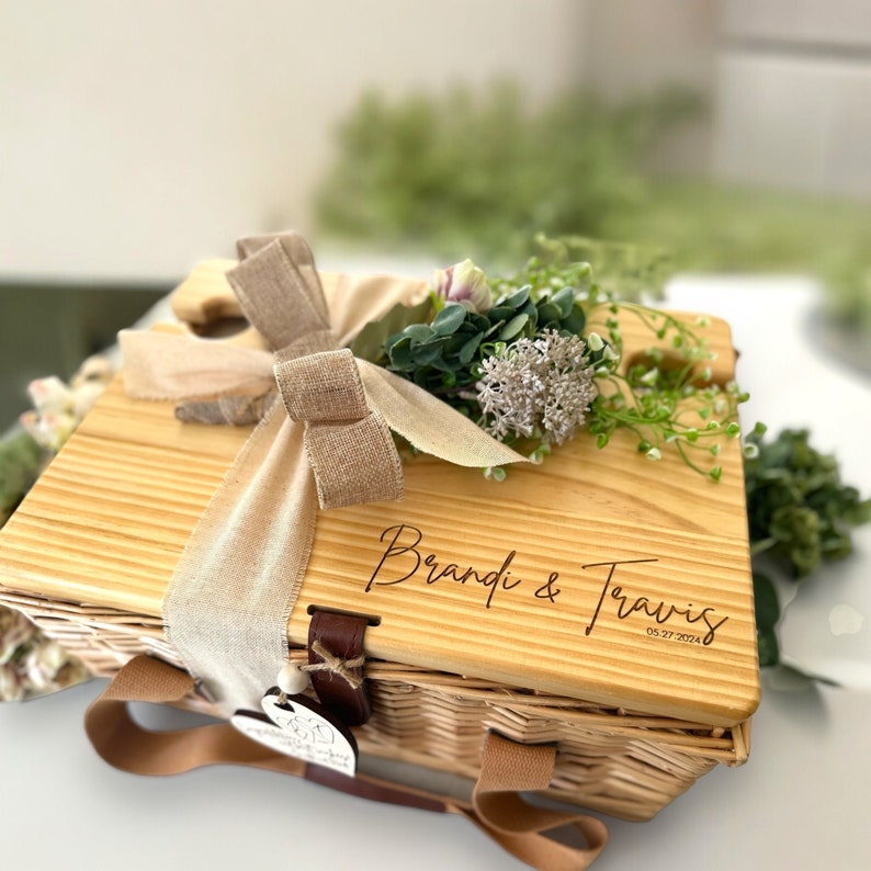 May include: A wooden picnic basket with a brown ribbon and a bouquet of flowers. The basket is personalized with the names "Brandi & Travis" and the date "05.27.2024".