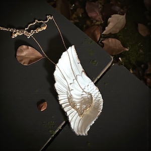 May include: A gold-colored necklace with an antler pendant rests on a white, wing-shaped dish. The necklace chain is delicate, and the antler design is detailed. The dish is placed on a dark surface with scattered leaves, suggesting a nature-inspired theme.