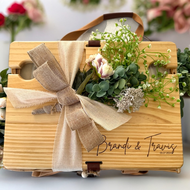 May include: A wooden picnic basket with a brown burlap ribbon and a bouquet of white and green flowers. The basket is engraved with the names "Brandi & Travis" and the date "05.27.2024".