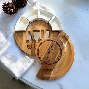 K&ouml;nnte beinhalten: Ein K&auml;sebrett-Set aus Holz mit rundem Design, mit kleinen wei&szlig;en Servierschalen und K&auml;semessern. Das Brett ist mit "The Sampson Family" graviert.