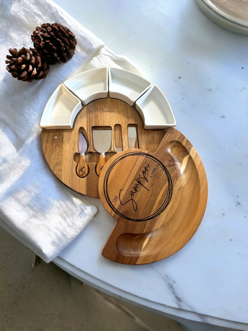 K&ouml;nnte beinhalten: Ein K&auml;sebrett-Set aus Holz mit rundem Design, mit kleinen wei&szlig;en Servierschalen und K&auml;semessern. Das Brett ist mit "The Sampson Family" graviert.