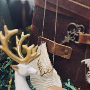 May include: A gold-toned antler necklace hangs in front of a brown leather journal. A white and gold deer figurine and a decorative key are also visible. The background includes green and white decorative paper.