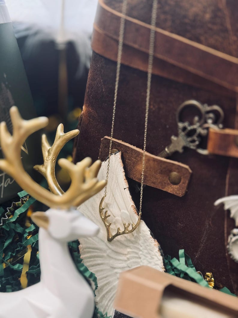 May include: A gold-toned antler necklace hangs in front of a brown leather journal. A white and gold deer figurine and a decorative key are also visible. The background includes green and white decorative paper.