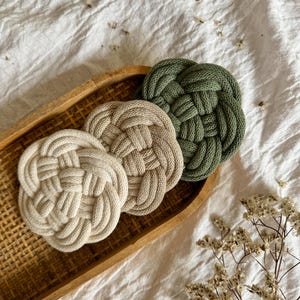 Könnte beinhalten: Drei runde, gewebte Untersetzer in einem Holztablett. Die Untersetzer sind in den Farbtönen Creme, Beige und Olivgrün gehalten. Die Untersetzer haben ein geknotetes Design. Das Tablett ist rechteckig und aus geflochtenem Holz. Getrocknete Blumen befinden sich in der unteren rechten Ecke.