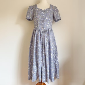 May include: A light blue floral print dress with short sleeves and a square neckline. The dress is buttoned down the front and has a fitted bodice that flows into a full skirt. The dress is displayed on a mannequin.