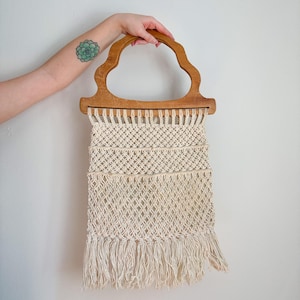 May include: A cream-colored macrame wall hanging with a wooden handle. The macrame design features a woven pattern with fringe at the bottom. The wooden handle has a unique, cloud-like shape.