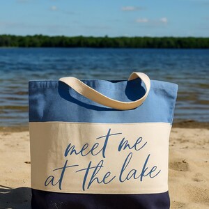 Op de afbeelding: Een blauwe en beige draagtas met de tekst "meet me at the lake" in een scriptlettertype. De tas heeft een lichtblauw bovenstuk, een beige middenstuk en een donkerblauw onderstuk. De tas staat op een zandstrand.