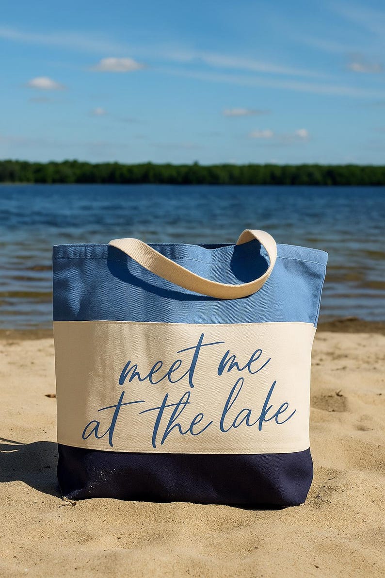 Op de afbeelding: Een blauwe en beige draagtas met de tekst "meet me at the lake" in een scriptlettertype. De tas heeft een lichtblauw bovenstuk, een beige middenstuk en een donkerblauw onderstuk. De tas staat op een zandstrand.