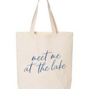 Op de afbeelding: Een cr&egrave;mekleurige draagtas met lange handvatten. De tas heeft de tekst "meet me at the lake" in een cursief, marineblauwe lettertype. De tas is gemaakt van een stevige stof, geschikt voor het dragen van spullen.