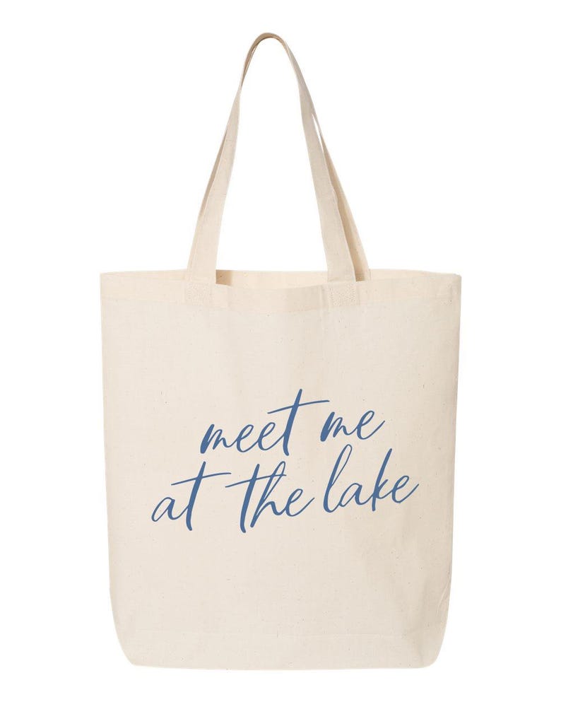 Op de afbeelding: Een cr&egrave;mekleurige draagtas met lange handvatten. De tas heeft de tekst "meet me at the lake" in een cursief, marineblauwe lettertype. De tas is gemaakt van een stevige stof, geschikt voor het dragen van spullen.