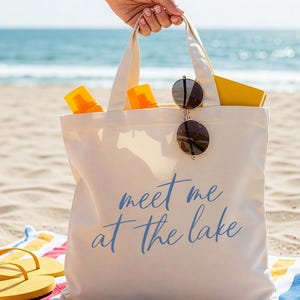 Op de afbeelding: Een cr&egrave;mekleurige draagtas met de tekst "meet me at the lake" in blauwe letters. De tas bevat een zonnebril, zonnebrandcr&egrave;me en een geel boek. De tas ligt op een gestreepte handdoek met gele slippers.