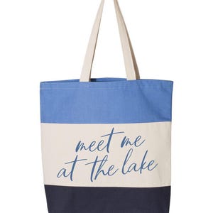 Op de afbeelding: Een canvas draagtas met een blauw, wit en marinekleurig blokontwerp. De tas heeft de tekst "meet me at the lake" in een cursief lettertype. De tas heeft cr&egrave;mekleurige handvatten.