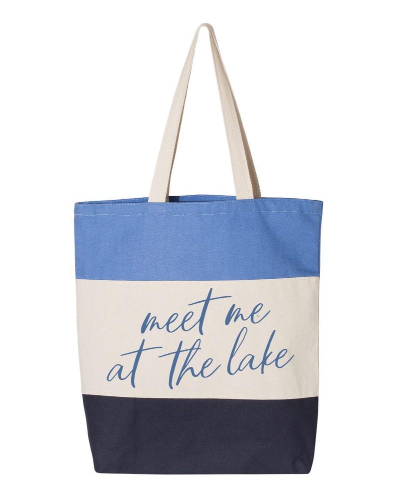 Op de afbeelding: Een canvas draagtas met een blauw, wit en marinekleurig blokontwerp. De tas heeft de tekst "meet me at the lake" in een cursief lettertype. De tas heeft cr&egrave;mekleurige handvatten.