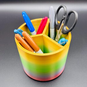 May include: A rainbow-coloured desk organiser with three compartments. The organiser holds a pair of scissors, a blue pen, a red pen, a brown pen, and two white markers.