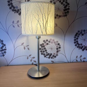 Peut inclure: Une lampe de table argentée avec un abat-jour cylindrique orné d'un motif de branches d'arbres blanches. La lampe est posée sur une surface en bois, avec du papier peint à motifs floraux en arrière-plan.