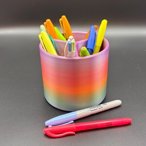 Peut inclure: Un organisateur de bureau arc-en-ciel avec trois compartiments contient une variété de stylos et de marqueurs. L'organisateur est en plastique et a une finition lisse et brillante. Deux marqueurs Sharpie sont posés sur une surface noire devant l'organisateur.