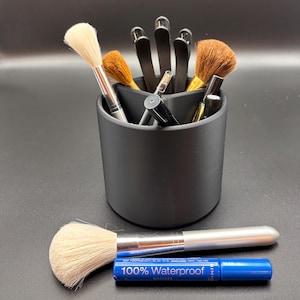 Puede incluir: Un soporte negro para brochas de maquillaje con tres compartimentos. El soporte está lleno de brochas de maquillaje, pinzas para el pelo y un tubo de máscara de pestañas. El tubo de máscara de pestañas es azul y dice "100% Waterproof Mascara".