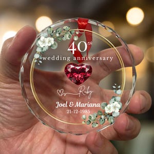 May include: A clear, round crystal ornament with a red ribbon. The ornament features a gold ring, the text "40th wedding anniversary", a ruby heart, and the names "Joel & Mariana" with the date "21-12-1985". Decorative floral elements are also present.