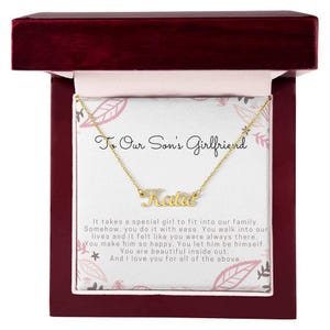 Könnte beinhalten: Eine goldfarbene Halskette mit dem Namen "Katie" in Schreibschrift, präsentiert in einer Geschenkbox. Die Box ist mit einem floralen Design versehen und enthält eine Nachricht an "Die Freundin unseres Sohnes".