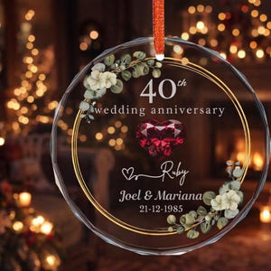 Puede incluir: Adorno redondo y transparente con borde dorado y cinta roja. Muestra el texto "40th wedding anniversary", un corazón de rubí y los nombres "Joel & Mariana" con la fecha "21-12-1985". Decoraciones florales adornan el diseño.
