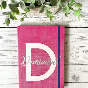 May include: A pink notebook with a white letter "D" and the name "Demianuk" written in silver lettering. The notebook has a blue elastic closure.