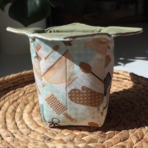 May include: A green and white fabric pot holder with a kitchen-themed pattern. The pot holder is folded and sitting on a woven straw mat.