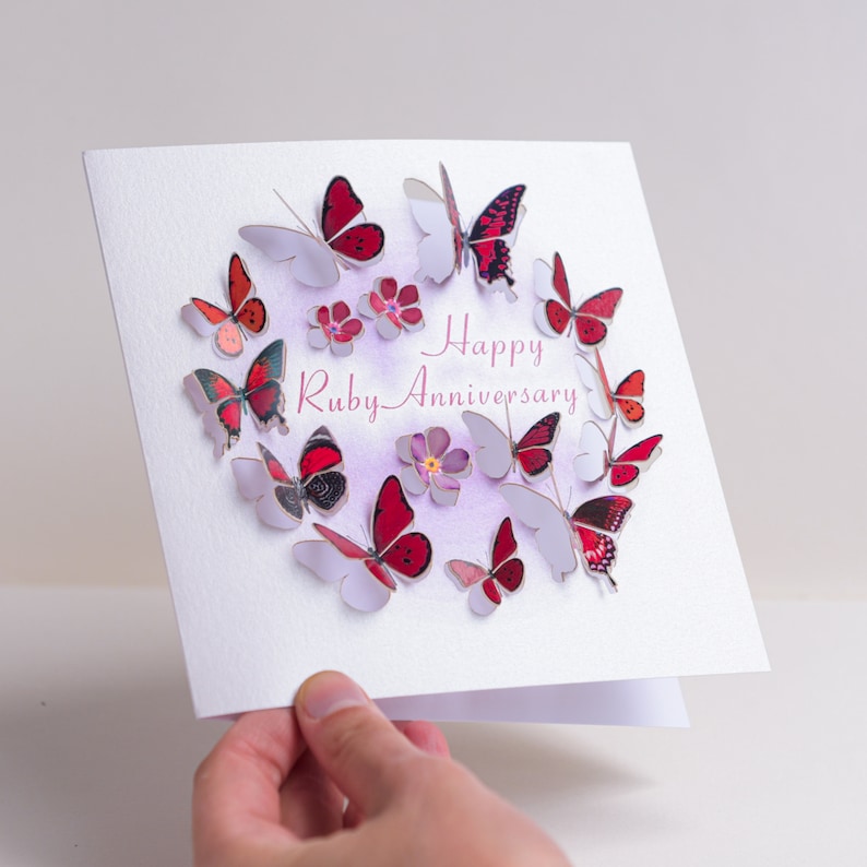 Peut inclure: Une carte blanche avec un fond aquarelle rose et violet. La carte a un cercle de papillons rouges et noirs avec le texte "Happy Ruby Anniversary" en rose.