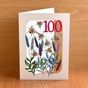 Puede incluir: Una tarjeta de felicitación de color crema con una ventana rectangular recortada. El diseño muestra varios abejorros ilustrados volando entre ramas de lavanda moradas y verdes. El número 100 está impreso en una fuente roja grande y negrita.