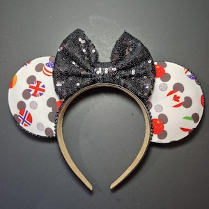 May include: Minnie Mouse ear headband with a black sequined bow. The ears feature a white background with gray Mickey Mouse heads and flags of various countries. The headband is beige with black beading.